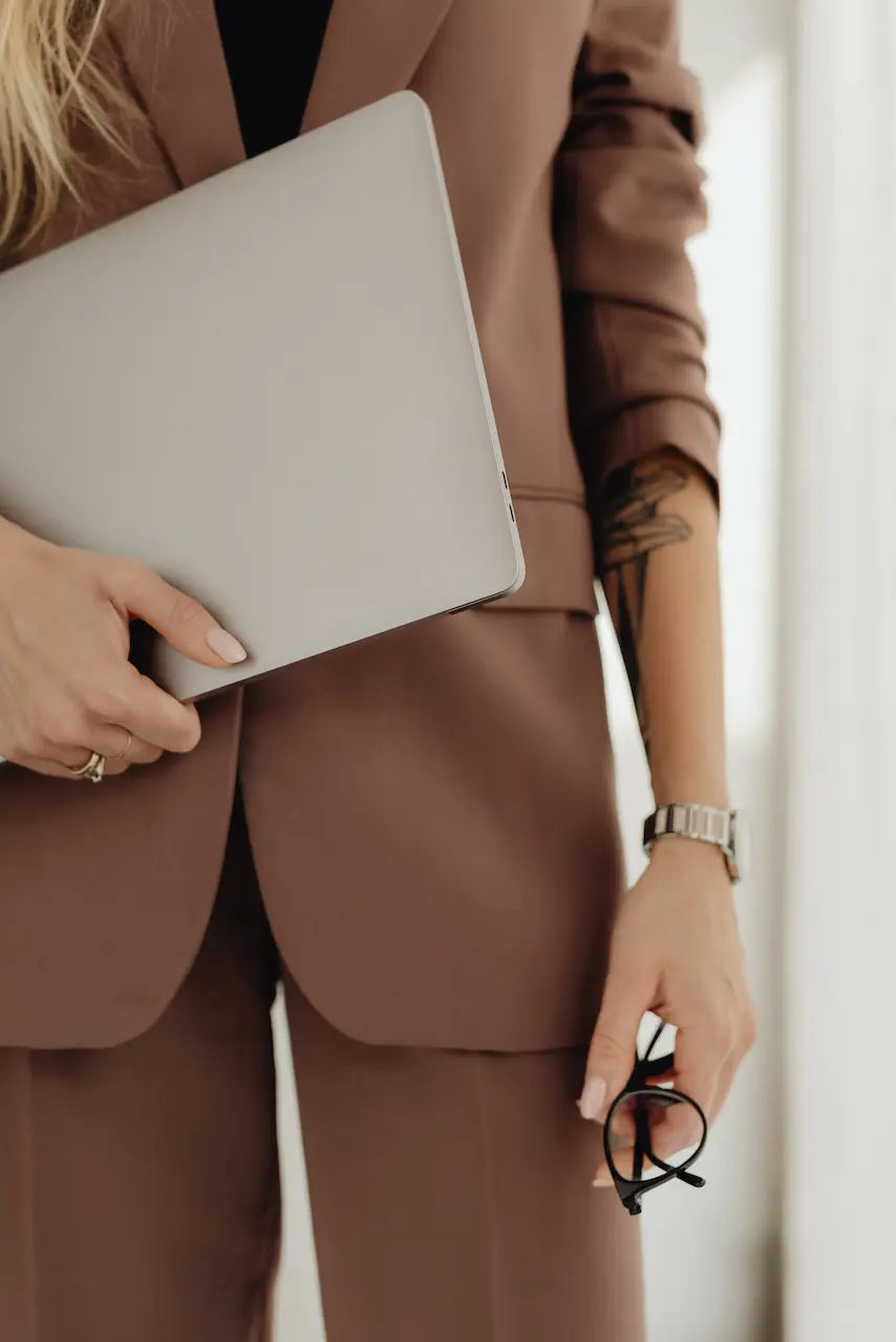 Femme en costume brun tenant un ordinateur sous son bras.